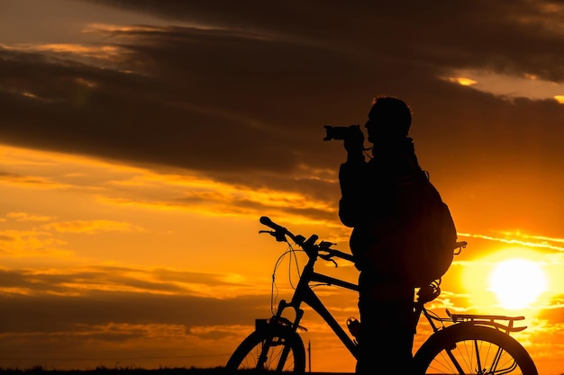 La silhouette d'un cycliste photographe masculin se tient sur la route et regarde au loin et admire le magnifique paysage au coucher du soleil cramoisi