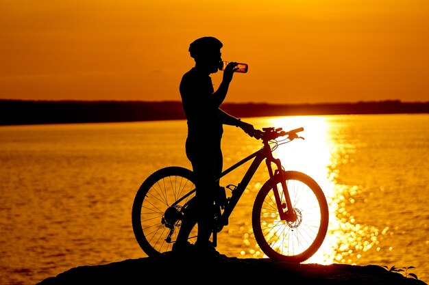 Silhouette de cyclistes au coucher du soleil. Les hommes boivent de l'eau. Rivière en arrière-plan. Concept de mode de vie.