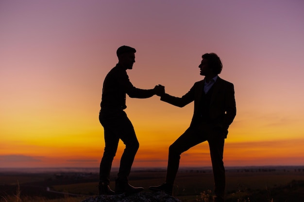 Photo silhouette de deux hommes dans le ciel coucher de soleil (ciel orange et rose) sur la colline rocheuse. deux messieurs se serrent la main