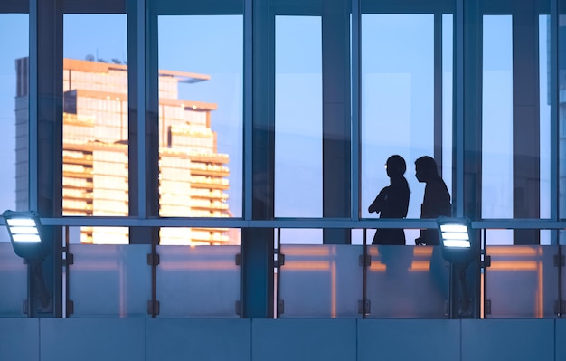 Silhouette de deux touristes marchant sur la passerelle en verre surélevée d'un bâtiment moderne au coucher du soleil