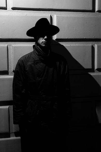 Silhouette dramatique sombre d'un homme dans un chapeau et un imperméable la nuit dans la rue dans un style noir