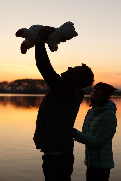 Silhouette du père tenant le petit bébé dans ses mains et de la mère derrière le coucher du soleil avec le lac