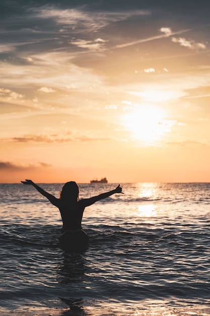Silhouette de femme assise sur une planche de surf sur la plage lève les bras avec un geste de surf devant le soleil au coucher du soleil