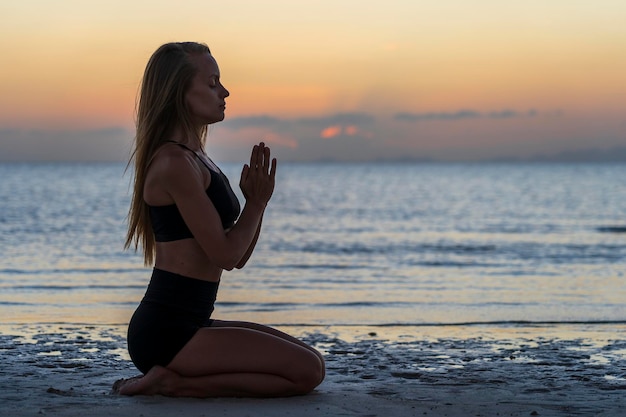 Silhouette de femme assise à la pose de yoga sur la plage tropicale pendant le coucher du soleil. Fille caucasienne pratiquant le yoga près de l'eau de mer.
