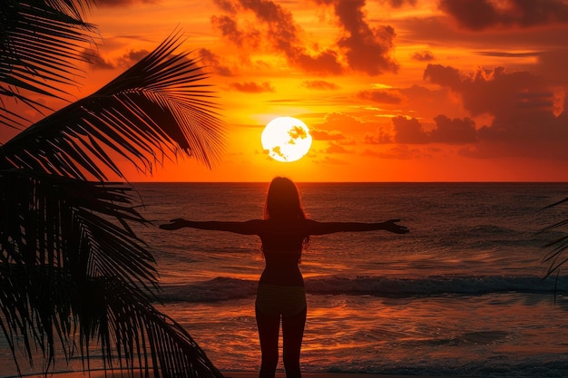 Silhouette d'une femme avec les bras tendus profitant d'une plage tropicale au coucher du soleil avec des palmiers et un ciel orange spectaculaire Concept de relaxation, de liberté et de paradis