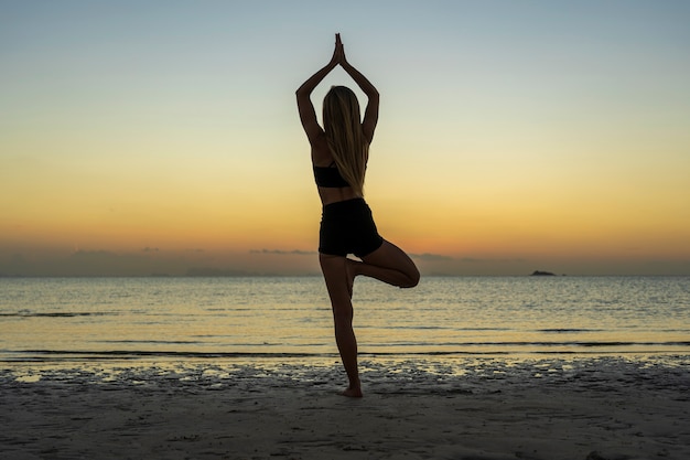 Photo silhouette de femme debout à la pose de yoga sur la plage tropicale pendant le coucher du soleil. fille caucasienne pratiquant le yoga près de l'eau de mer