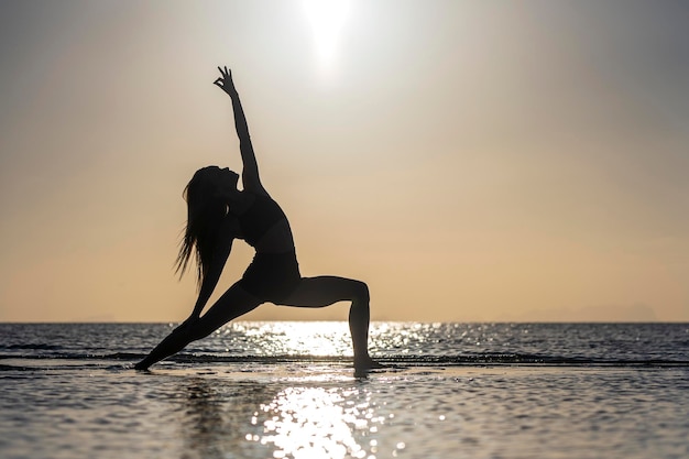 Silhouette de femme debout à la pose de yoga sur la plage tropicale pendant le coucher du soleil. Fille caucasienne pratiquant le yoga près de l'eau de mer