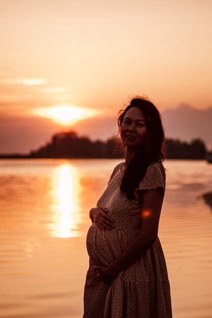 Silhouette de femme enceinte souriante heureuse jeune femme en robe légère profite des rayons du soleil couchant...