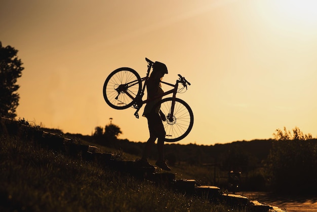 Silhouette d'une femme portant un vélo contre le ciel du soir