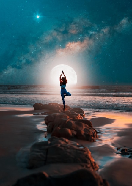 Silhouette d'une femme en pose de yoga sur des rochers sur la plage la nuit avec la lune et la voie lactée