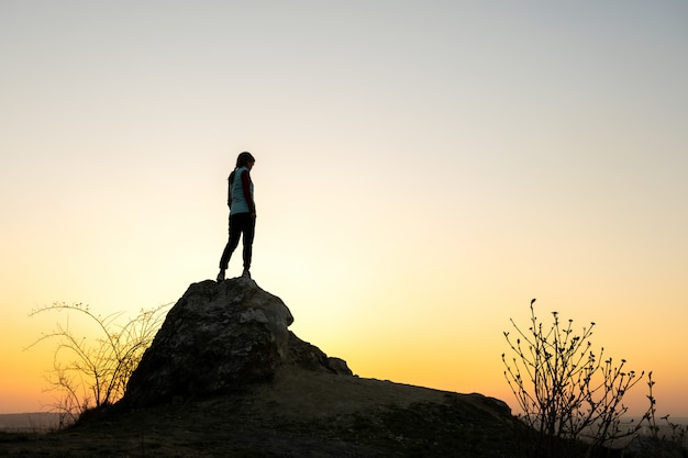 Silhouette d'une femme randonneur debout seul sur une grosse pierre au coucher du soleil dans les montagnes. Touriste sur haut rocher dans la nature du soir. Concept de tourisme, de voyage et de mode de vie sain.