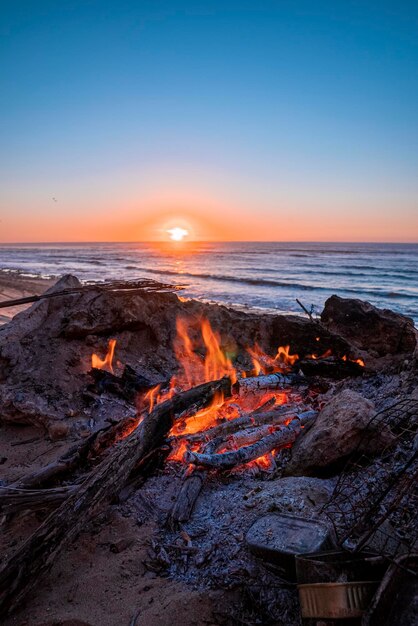 Silhouette de feu avec du bois de chauffage brûlant contre un beau soleil se couchant à la plage