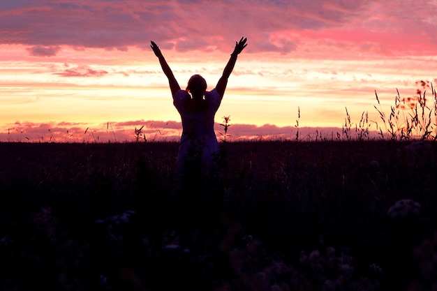 Silhouette d'une fille dans un champ avec les mains levées contre le ciel coucher de soleil