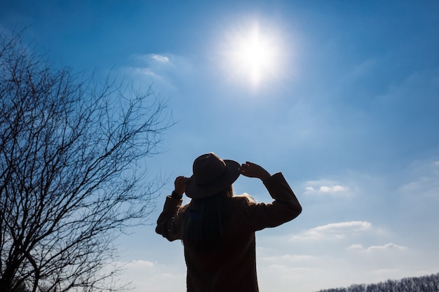 Silhouette d'une fille dans un chapeau et un manteau sur un fond de ciel avec des nuages et le soleil.