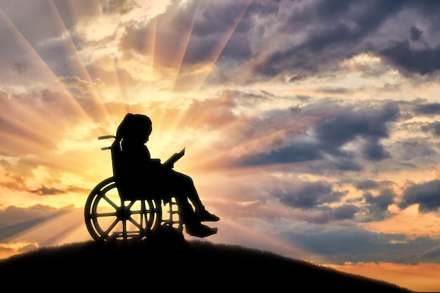 Silhouette d'une fille enfant handicapée assise dans un fauteuil roulant, lisant un livre sur un fond de coucher de soleil sur une colline. La vie des enfants handicapés
