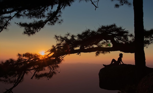 Une silhouette de fille est assise sur un rocher de falaise avec un ciel crépusculaire au coucher du soleil