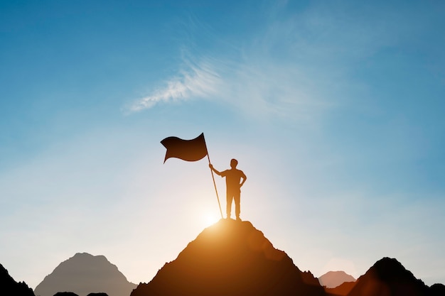 Silhouette d'homme d'affaires tenant le drapeau au sommet de la montagne avec plus de ciel bleu et la lumière du soleil.