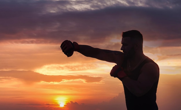 Silhouette d'homme athlétique combattant avec des gants de boxe posant combat et biceps au fond du coucher du soleil