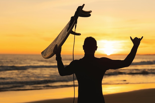 Silhouette d'un homme athlétique tenant une planche de surf montrant shaka au coucher du soleil