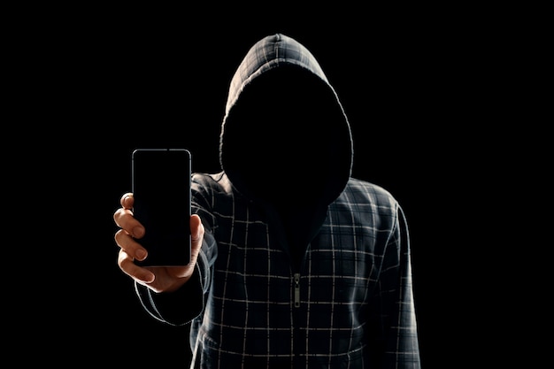 Silhouette d'un homme dans une cagoule sur un fond noir son visage n'est pas visible le pirate informatique tient le téléphone dans ses mains