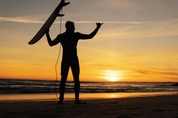 Silhouette d'homme debout sur le bord de la mer tenant une planche de surf et montrant shaka au coucher du soleil