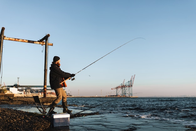 Silhouette d'homme pêcheur portant un manteau, tenant une tige, pêchant à la plage