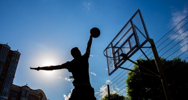 Silhouette d'un homme qui lance une balle dans un panier de basket dans la rue contre un ciel avec des nuages de couleur noir et blanc