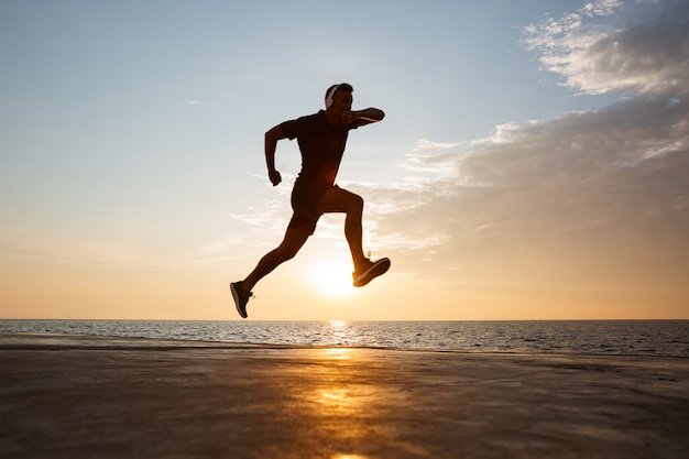 Silhouette d'homme sportif actif 30 s sauter et courir le long de la jetée au bord de la mer, et écouter de la musique via des écouteurs sans fil pendant le lever du soleil