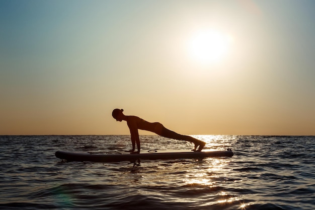 Silhouette de jeune belle fille pratiquant le yoga sur planche de surf en mer au lever du soleil.