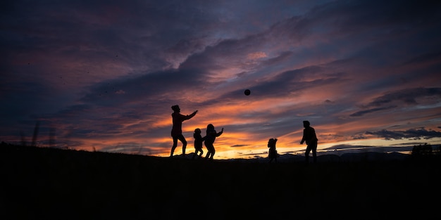 Silhouette d'une jeune famille de cinq personnes jouant avec un ballon à l'extérieur sous un beau ciel dramatique au coucher du soleil.