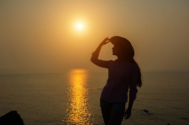 Silhouette de jeune femme au chapeau contre le coucher de soleil sur la mer