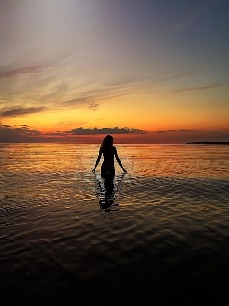 silhouette de jeune femme au coucher du soleil orange dans la vague d'eau de mer et ciel nuageux rose