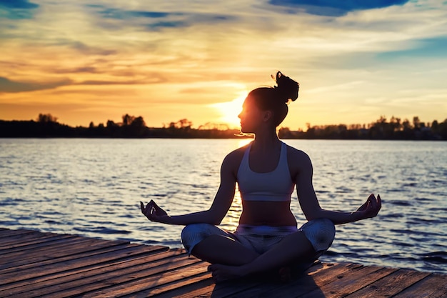 Silhouette d'une jeune femme faisant des exercices de yoga sur la plage du lac au coucher du soleil