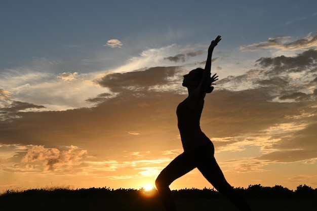 Silhouette de jeune femme mince pratiquant la pose de guerrier de yoga, coucher du soleil à l'arrière-plan