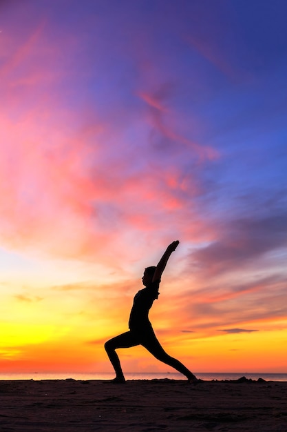 Silhouette jeune femme pratiquant le yoga sur la plage au coucher du soleil.