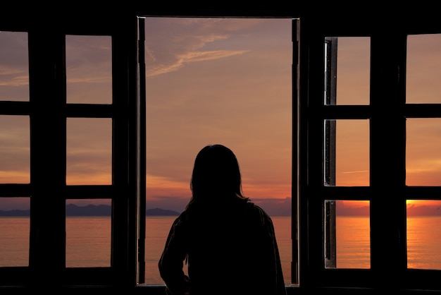 La silhouette d&#39;une jeune femme regardant le coucher du soleil par la fenêtre avec vue sur la mer.