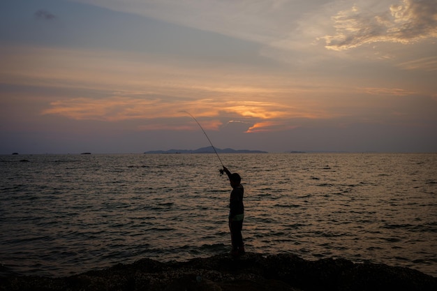 Silhouette jeune garçon pêchant au bord de la mer contre le coucher du soleil