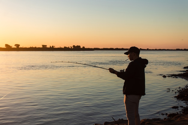 Silhouette d'un jeune homme fumant une cigarette et pêchant dans la rivière.