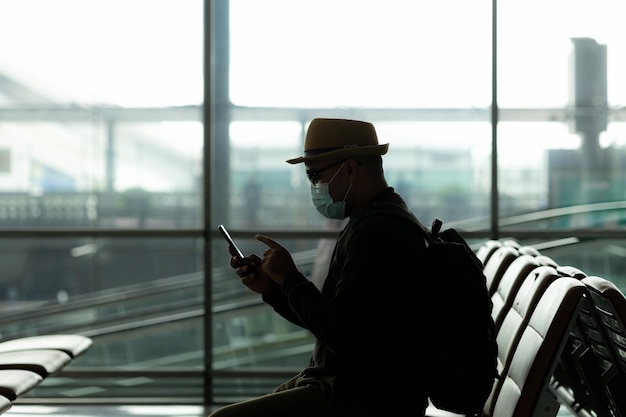 silhouette d'un jeune sac à dos de voyage à l'aide d'un smartphone debout dans le bâtiment d'arrivée de départ des passagers à l'aéroport. Il voyage seul en gare.