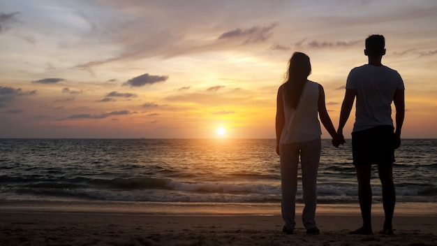 Silhouette de jeunes marchant sur la plage à l'eau de mer sans limites se joignant les mains au coucher du soleil orange vue arrière