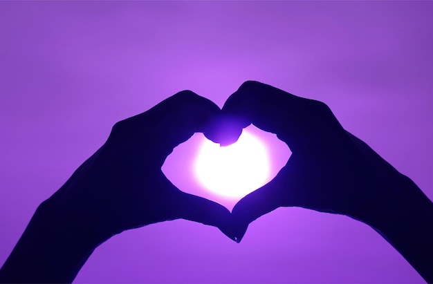 Silhouette de la main de la femme posant le signe LOVE HEART contre le soleil brillant sur un ciel violet vibrant