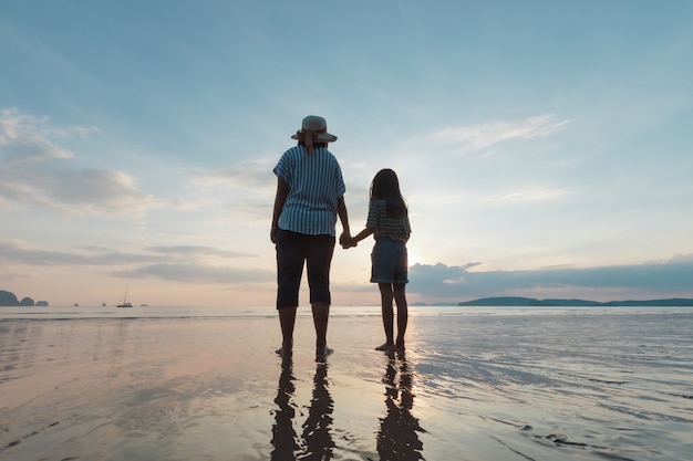 Silhouette de mère asiatique et fille tenant la main ensemble debout sur la plage en regardant la belle mer et le ciel au moment du coucher du soleil.