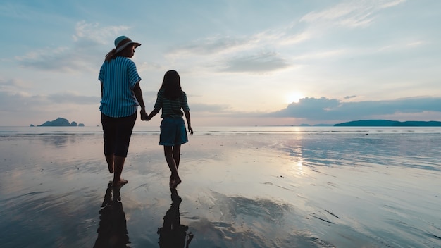 Silhouette de mère et fille asiatique tenant la main et marchant sur la plage ensemble au moment du coucher du soleil avec une belle mer et ciel. Profitez de la famille avec le concept de la nature.