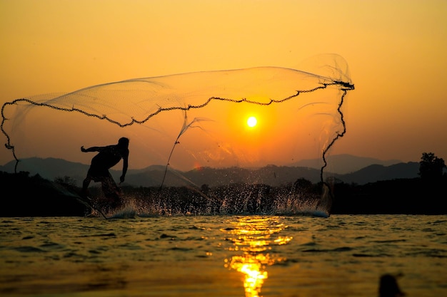 Silhouette d'un pêcheur jetant un filet dans la rivière contre un ciel orange