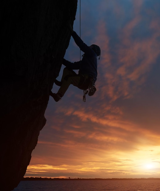 Silhouette de personne athlétique dans le harnais de sécurité Grimpeur sur corde escalade sur falaise difficile au-dessus de l'eau au coucher du soleil