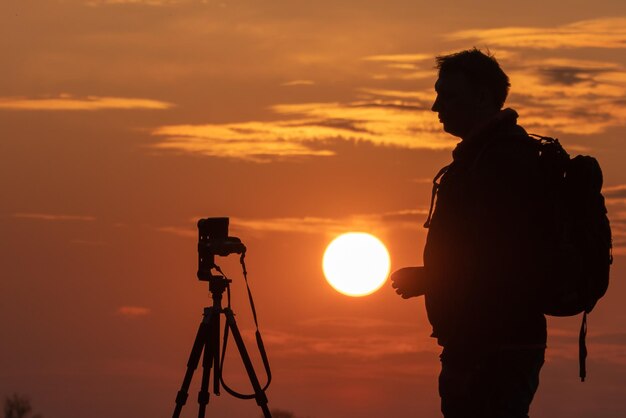 La silhouette d'une personne sur fond de soleil et de beau ciel Le contour d'un homme debout à côté d'un trépied et d'un appareil photo sur fond de coucher de soleil