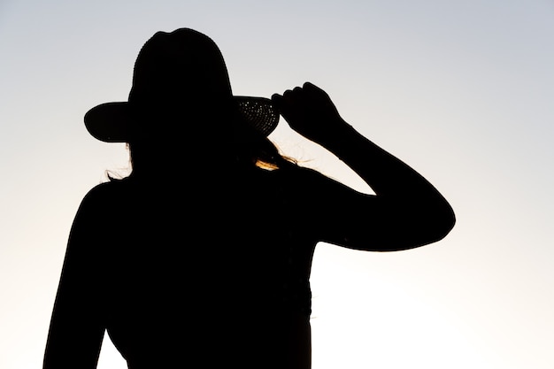 Silhouette rétroéclairée d'une femme tenant un chapeau d'une main.