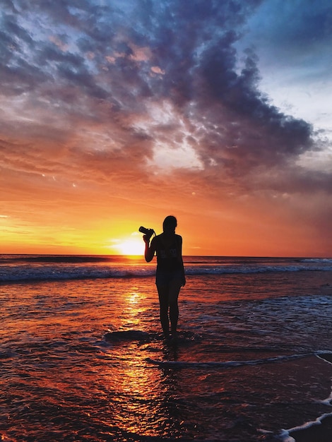 Silhouette sombre d'une femme avec un appareil photo sur l'océan dans un coucher de soleil orange