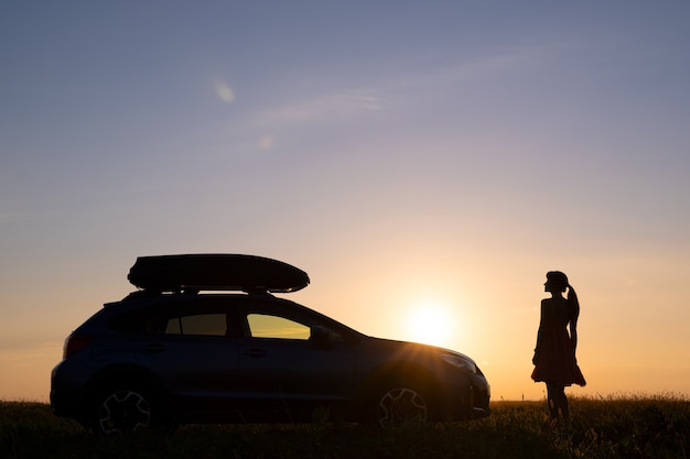 Silhouette sombre d'une femme solitaire se relaxant près de sa voiture sur une prairie herbeuse, profitant de la vue sur le lever du soleil coloré.