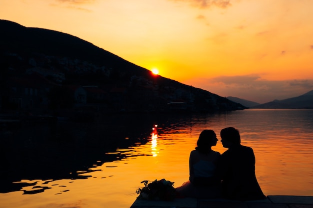 Des silhouettes de couple s'assoient sur la jetée au-dessus de l'eau sur fond de montagnes et de coucher de soleil